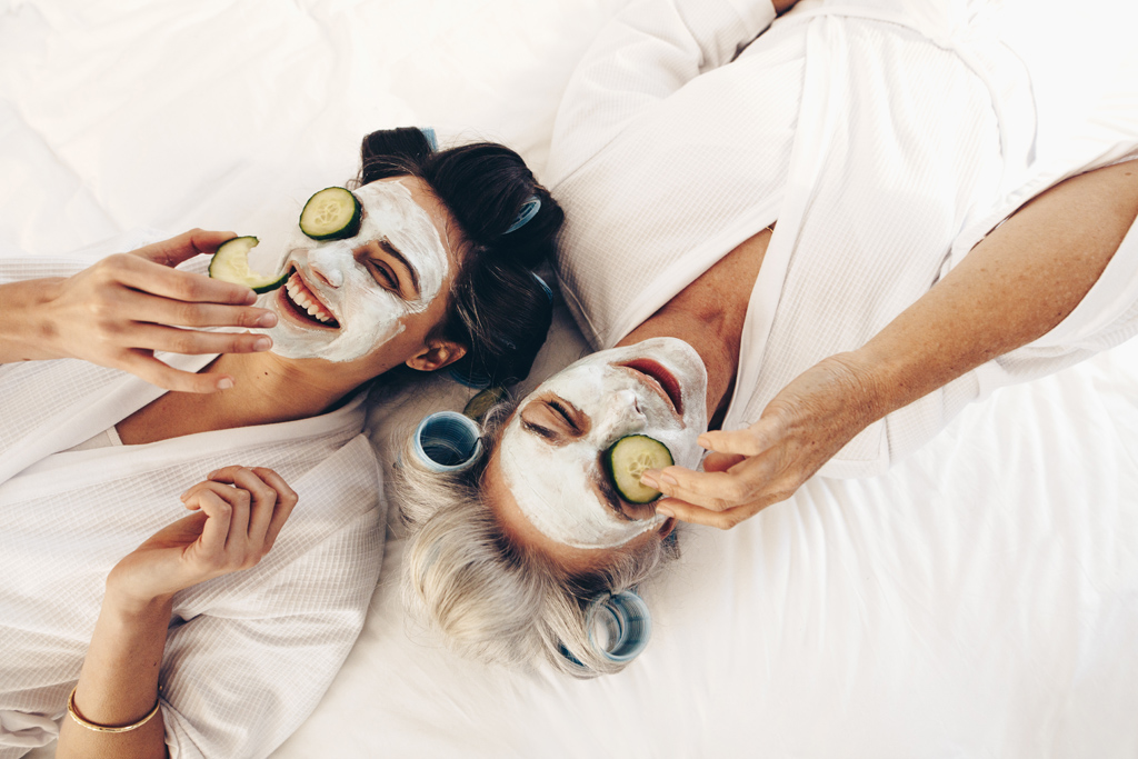 Two women in bathrobes lying on bed wearing beauty face pack with cucumber slices. Mother and daughter relaxing on bed wearing face pack and roller curls on hair.