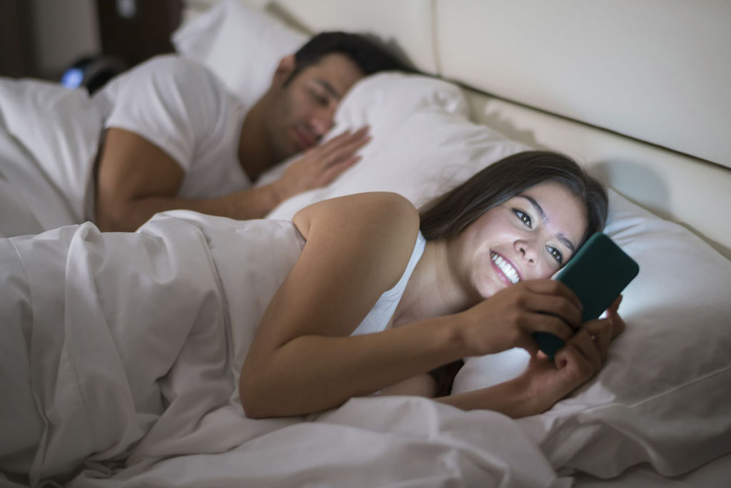 Woman texting or social networking on her smart phone in bed while her husband is sleeping